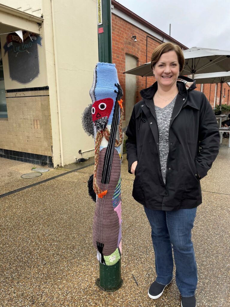 Beth standing next to a small street bollard that has been covered with a knitted yarn cover designed to look like a person playing a guitar. The background show some footpath and buildings in Chiltern.